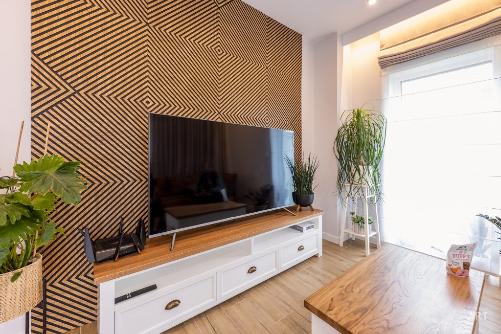 Nowoczesny salon z telewizorem zamontowanym na ścianie z geometrycznym wzorem w paski oraz białą szafką RTV z drewnianym blatem. W pomieszczeniu znajdują się rośliny doniczkowe, drewniany stolik kawowy i duże okno z roletą.