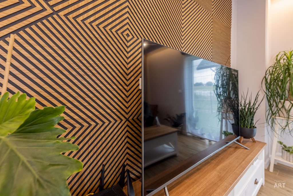 Telewizor umieszczony na drewnianej szafce stoi przy ścianie z geometrycznym wzorem w paski tworzącym efekt optyczny. Obok telewizora znajdują się doniczki z zielonymi roślinami, a w lewym dolnym rogu widać liście dużej rośliny.