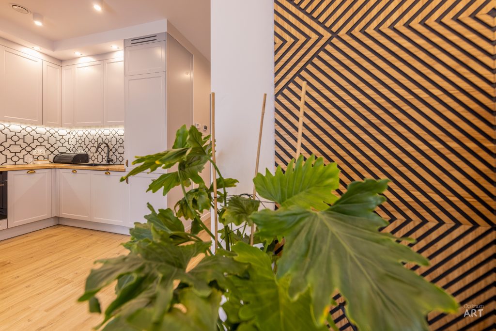Nowoczesna kuchnia z białymi szafkami i geometryczną tapetą na ścianie w tle. Na pierwszym planie widać dużą roślinę doniczkową z szerokimi zielonymi liśćmi.
