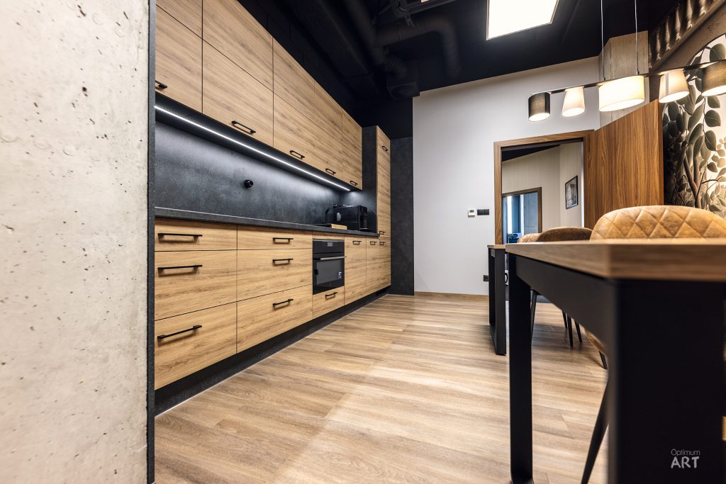 Nowoczesna kuchnia w stylu industrialnym z drewnianymi frontami szafek, czarnymi elementami i betonową ścianą. W tle widać jadalnię ze stołem i krzesłami oraz otwarte przejście do kolejnego pomieszczenia.