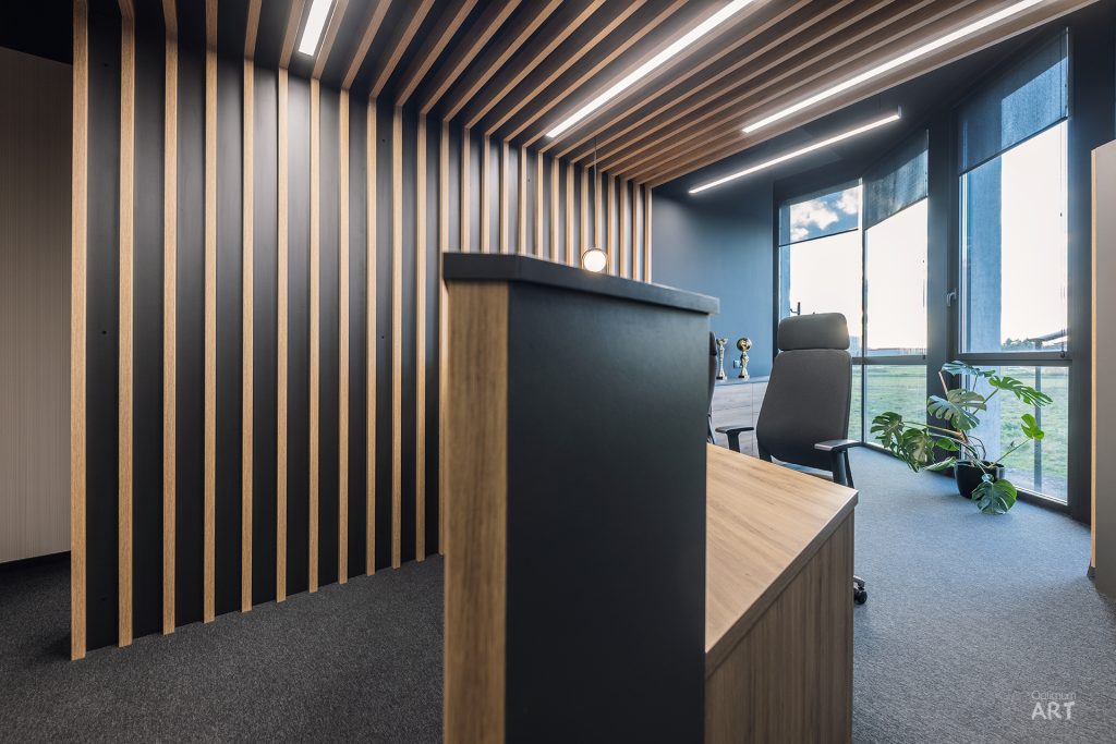 Nowoczesne biuro z drewnianymi lamelami na ścianie i suficie, w którym znajduje się duże biurko z czarnym krzesłem obrotowym. Pomieszczenie ma duże okna z widokiem na zewnątrz i roślinę doniczkową przy oknie.