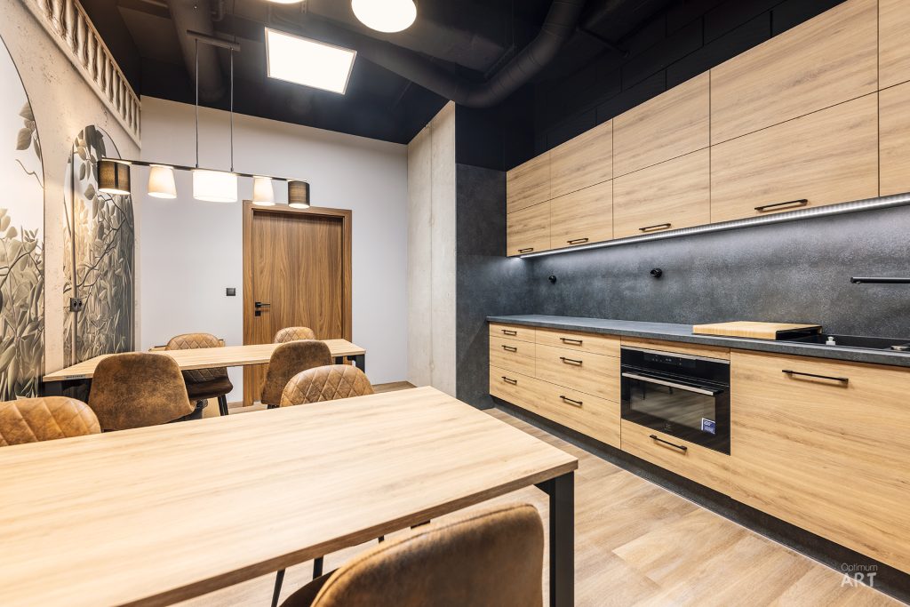Nowoczesna kuchnia z jasnymi drewnianymi szafkami i ciemnoszarym blatem, połączona z jadalnią ze stołami i brązowymi krzesłami. Wnętrze ma czarny sufit z belkami, jasne ściany i podłogę w kolorze drewna.