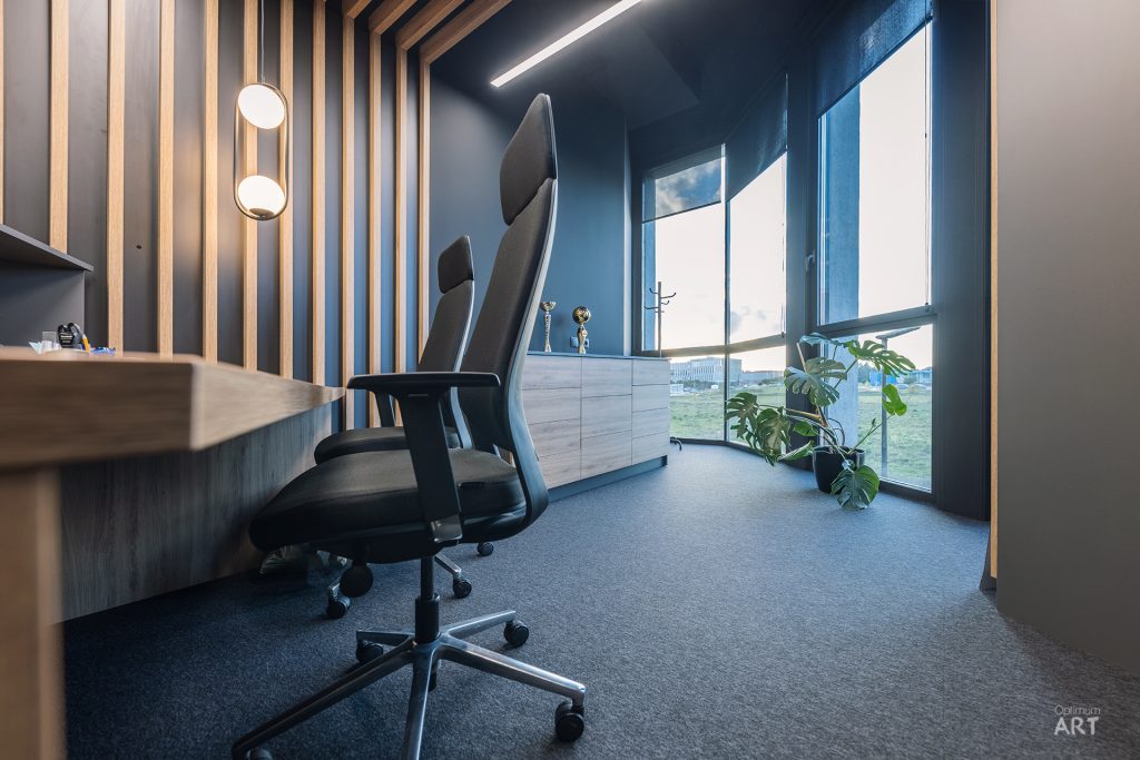 Nowoczesne biuro z czarnym fotelem obrotowym przy drewnianym biurku, z pionowymi drewnianymi lamelami na ścianie i dużymi oknami z widokiem na zewnątrz. Na podłodze leży ciemnoszary dywan, a przy oknie stoi roślina doniczkowa.