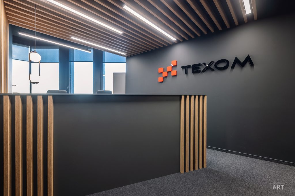 Nowoczesne biuro recepcyjne z drewnianym kontuarem i sufitem z drewnianych listew, na ciemnej ścianie widnieje logo "TEXOM" z pomarańczowymi kwadratami. W tle znajdują się duże okna i krzesła w strefie oczekiwania.