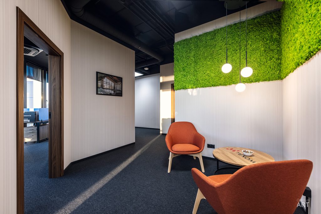 Nowoczesne wnętrze biurowe z dwoma pomarańczowymi fotelami ustawionymi wokół okrągłego stolika, na tle ściany pokrytej zieloną roślinnością. Po lewej stronie widoczne są drzwi prowadzące do sąsiedniego pomieszczenia, a nad strefą wypoczynkową wiszą białe lampy kuliste.