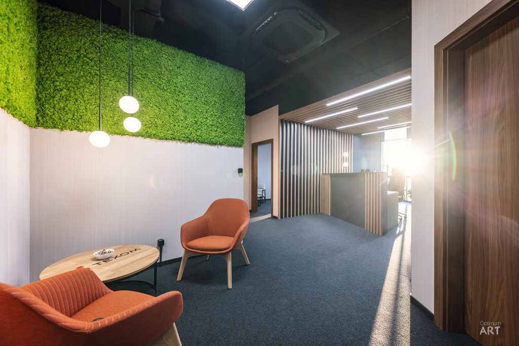 Nowoczesne wnętrze biurowe z zieloną ścianą z mchu, dwoma pomarańczowymi fotelami i okrągłym stolikiem. Przestrzeń ma niebieską wykładzinę, drewniane elementy wykończeniowe i wiszące białe lampy.