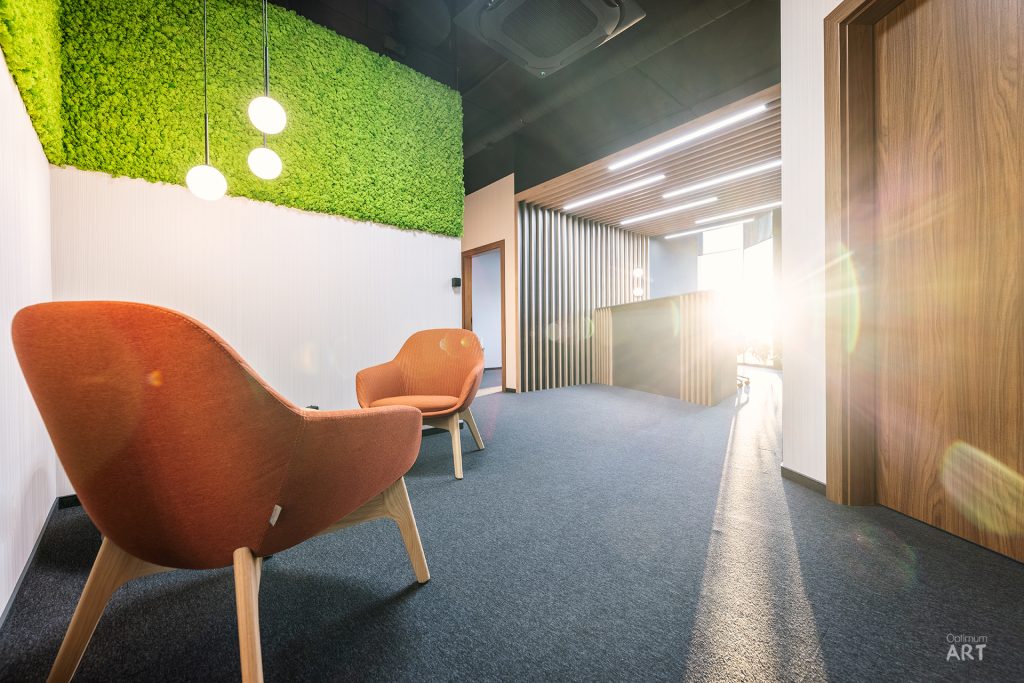 Nowoczesne wnętrze biurowe z dwoma pomarańczowymi fotelami na niebieskim dywanie, zieloną ścianą z mchu na suficie i drewnianymi panelami. Przestrzeń ma industrialny styl z czarnym sufitem, białymi ścianami i naturalnym oświetleniem wpadającym przez okno.