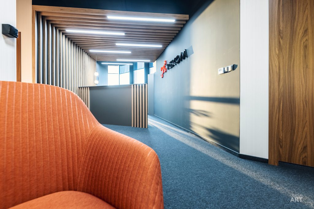 Nowoczesne wnętrze biurowe z pomarańczowym fotelem na pierwszym planie, drewnianym sufitem z belkami oraz logo firmy na ścianie. Przestrzeń charakteryzuje się niebieską wykładziną, jasnymi ścianami i współczesnym designem.