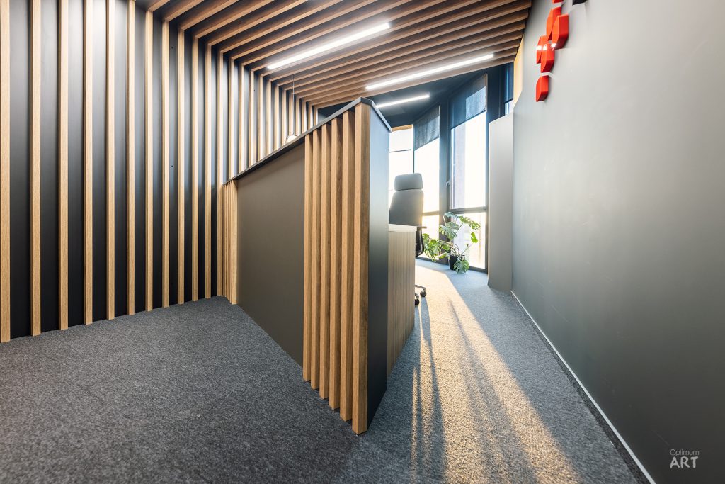 Nowoczesne wnętrze biurowe z charakterystyczną drewnianą lamelową ścianą i sufitem, które tworzą tunel optyczny prowadzący w głąb korytarza. Na prawej ścianie widoczny jest czerwony napis, a podłoga wykładzina jest szarym wykładzinem dywanowym.