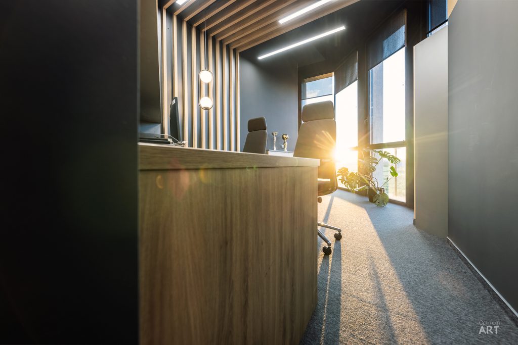 Nowoczesne biuro z drewnianym biurkiem, czarnym krzesłem obrotowym i charakterystycznym sufitem z drewnianych listew. Przez duże okno wpada jasne światło słoneczne, oświetlając przestrzeń z szarą wykładziną i rośliną w donicy.