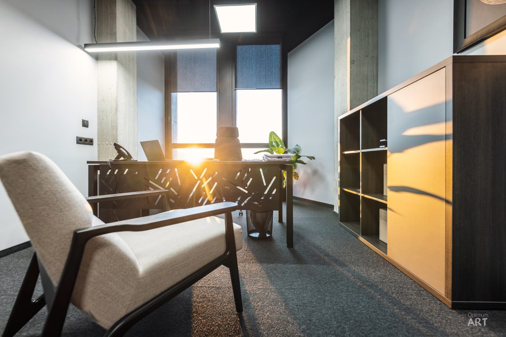 Nowoczesne biuro z drewnianym biurkiem przy oknie, na którym stoi laptop i roślina, oraz beżowym fotelem na pierwszym planie. Wnętrze ma ciemną kolorystykę z drewnianymi meblami i regałami, a przez okno wpadają promienie słońca.