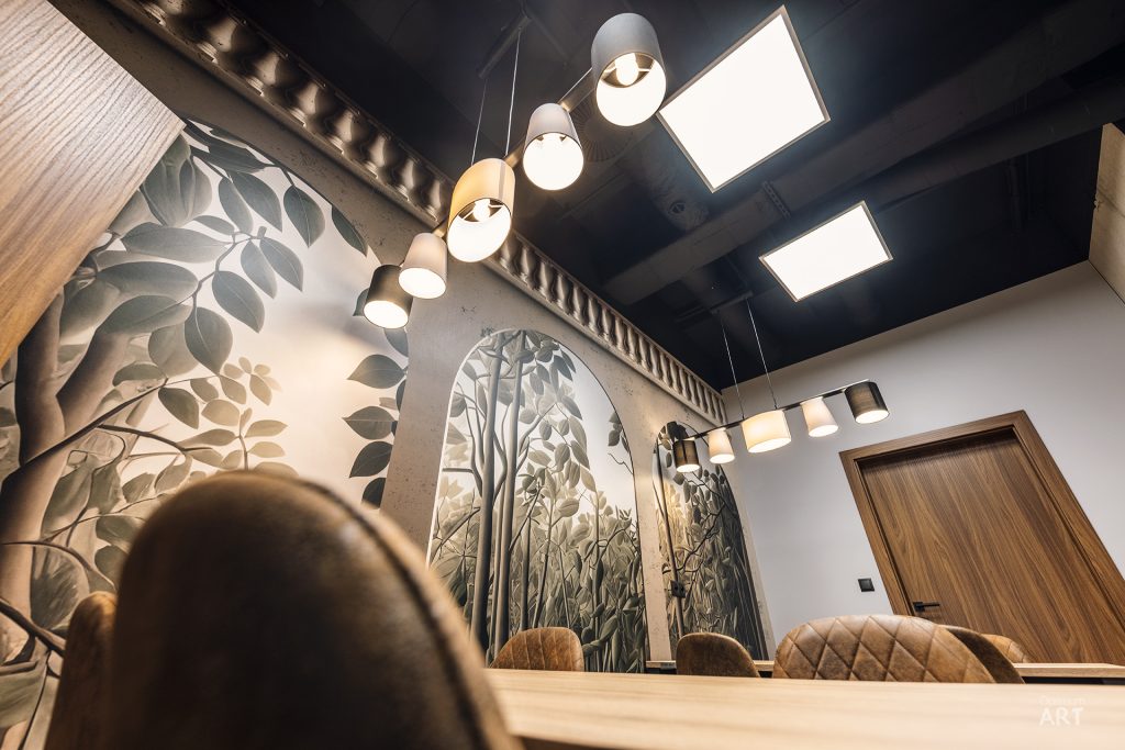 Nowoczesne wnętrze restauracji lub kawiarni z dekoracyjną ścianą pokrytą roślinnym motywem i łukowatymi niszami. Na suficie wiszą designerskie lampy wzdłuż drewnianej belki, a w pomieszczeniu widoczne są brązowe fotele przy drewnianym stole.