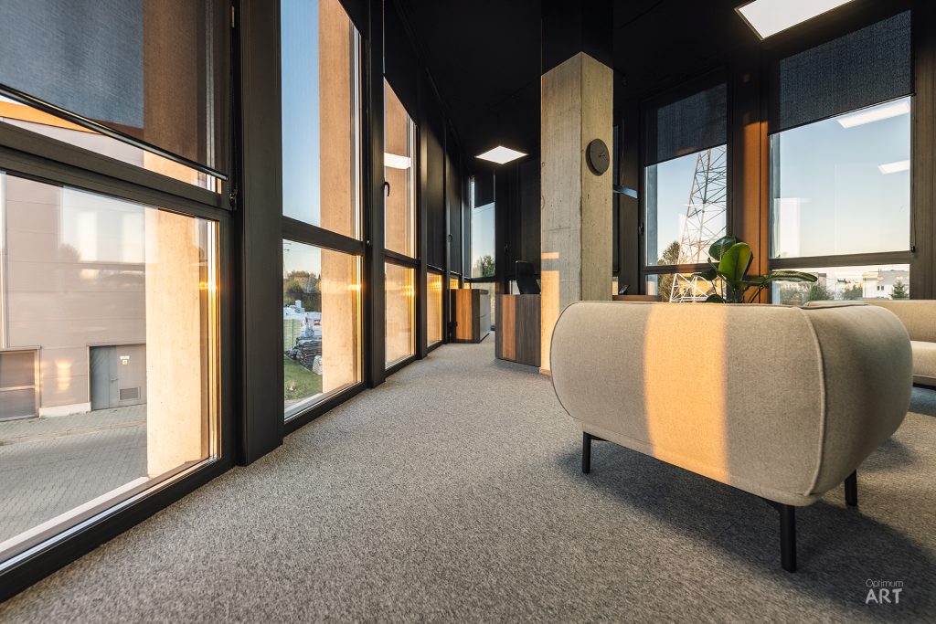 Nowoczesne wnętrze z dużymi oknami od podłogi do sufitu i beżowym fotelem na szarym dywanie. Przestrzeń ma ciemny sufit, jasne ściany i oferuje widok na zewnętrzne budynki przez panoramiczne okna.