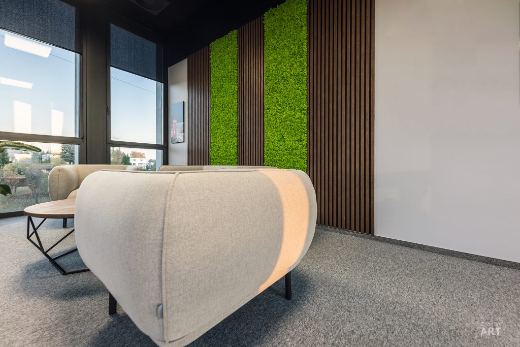Nowoczesne wnętrze biurowe z beżowym fotelem na pierwszym planie, ścianą z zielonymi panelami roślinnymi i drewnianymi lamelami w tle. Po lewej stronie znajdują się duże okna z widokiem na miasto, a na podłodze leży szary dywan.