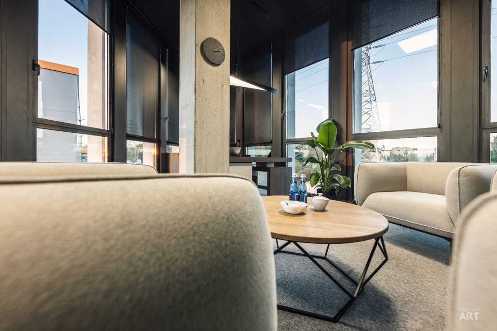 Nowoczesny salon z beżowymi fotelami ustawionymi wokół okrągłego drewnianego stolika kawowego, z dużymi oknami i betonową kolumną. Na stoliku stoją białe miseczki i roślina w doniczce, a w tle widoczny jest zegar na ścianie.