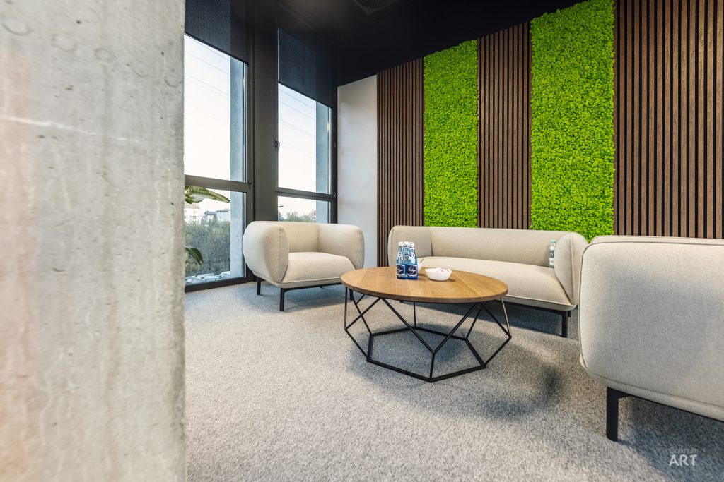 Nowoczesne wnętrze biurowe z beżowymi fotelami ustawionymi wokół geometrycznego stolika kawowego z drewnianym blatem. W tle widoczna ściana z zielonymi panelami roślinnymi i drewnianymi lamelami oraz duże okna wpuszczające naturalne światło.