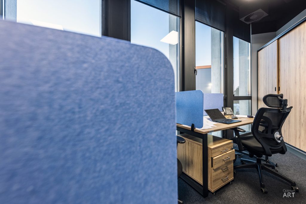 Nowoczesne biuro z drewnianym biurkiem, czarnym fotelem ergonomicznym i dużymi oknami. Na pierwszym planie widoczny niebieski panel akustyczny oddzielający przestrzeń biurową.