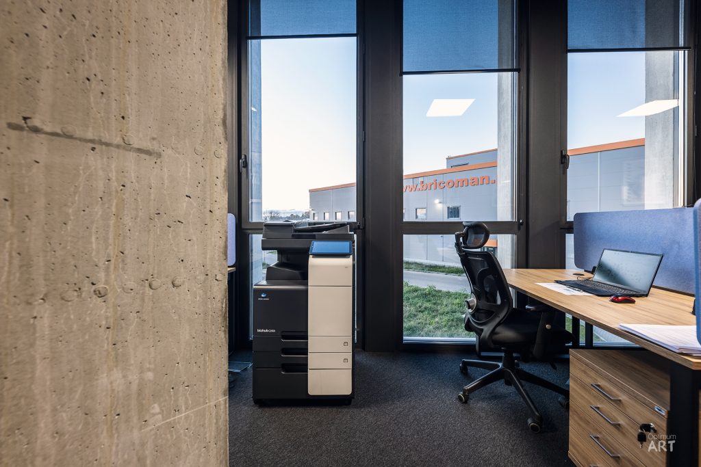 Nowoczesne biuro z drewnianym biurkiem, czarnym krzesłem obrotowym i laptopem, umieszczone przy dużych oknach z widokiem na budynki. Po lewej stronie znajduje się betonowa ściana, a na podłodze ciemna wykładzina dywanowa.