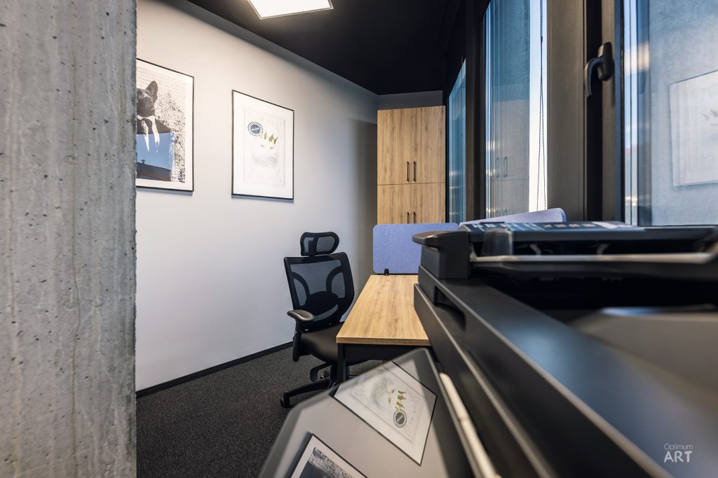Małe nowoczesne biuro z czarnym krzesłem biurowym przy drewnianym biurku, betonową ścianą po lewej stronie i dwoma oprawionymi obrazami na białej ścianie. W pomieszczeniu znajduje się również drewniana szafka i duże okno wpuszczające naturalne światło.