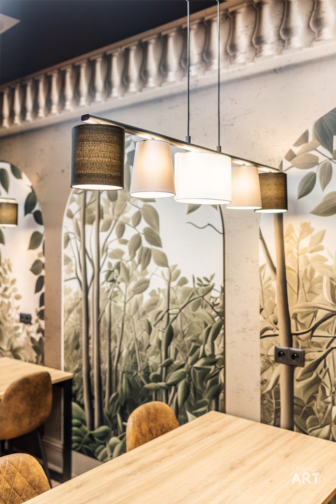 Wnętrze restauracji z drewnianym stołem, brązowymi krzesłami i wiszącą lampą z kolorowymi abażurami. Na ścianie widoczna jest tapeta z motywem roślinnym przedstawiającym zielone liście i gałęzie.