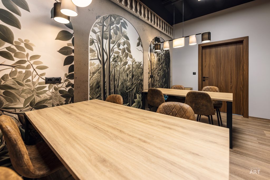 Nowoczesna sala konferencyjna z drewnianymi stołami i brązowymi krzesłami z pikowanymi oparciami. Na ścianach znajdują się dekoracyjne malowidła przedstawiające rośliny i liście w odcieniach zieleni i beżu.