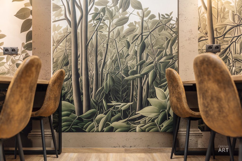 Wnętrze z dwoma drewnianymi krzesłami ustawionymi przy ścianie z dużą fototapetą przedstawiającą las z bambusami i zielonymi liśćmi. Na ścianie widoczne są gniazdka elektryczne, a całość utrzymana w naturalnych, ziemistych kolorach.