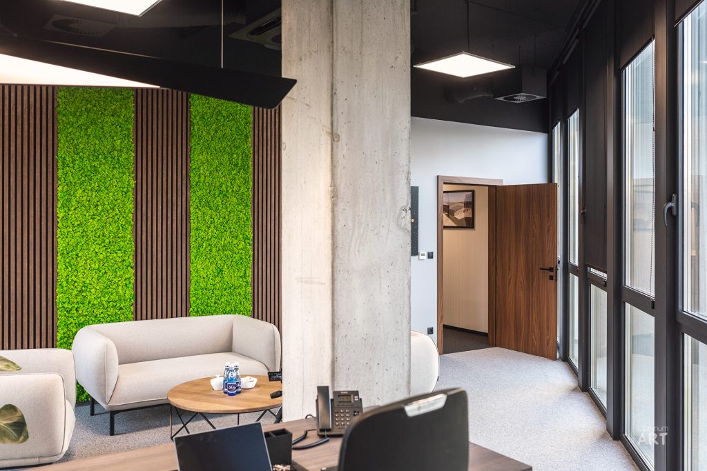 Nowoczesne wnętrze biurowe z betonowym słupem, beżowymi fotelami przy okrągłym stoliku i charakterystyczną ścianą z pionowymi drewnianymi lamelami przeplatanymi zielonym mchem dekoracyjnym. Przestrzeń ma duże okna, czarny sufit z widocznymi instalacjami oraz prowadzi do dalszych pomieszczeń z drewnianymi drzwiami.