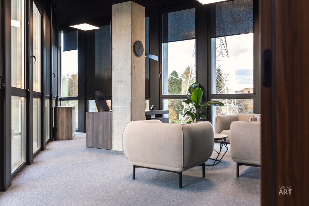 Nowoczesne wnętrze biurowe z dużymi oknami, betonową kolumną i beżowymi fotelami na szarym dywanie. W pomieszczeniu znajdują się także drewniane meble, roślina doniczkowa i czarne elementy wykończeniowe.