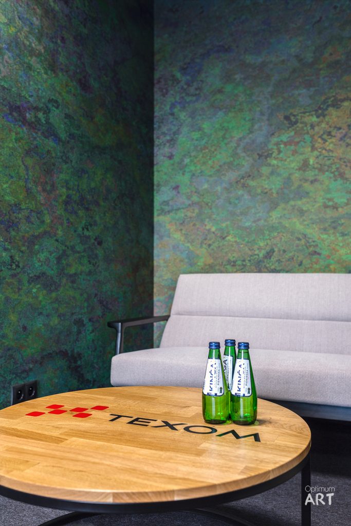 Na okrągłym drewnianym stoliku z napisem "HOTEL" stoją cztery zielone butelki wody. W tle widać jasną sofę i ścianę w odcieniach zieleni i błękitu.