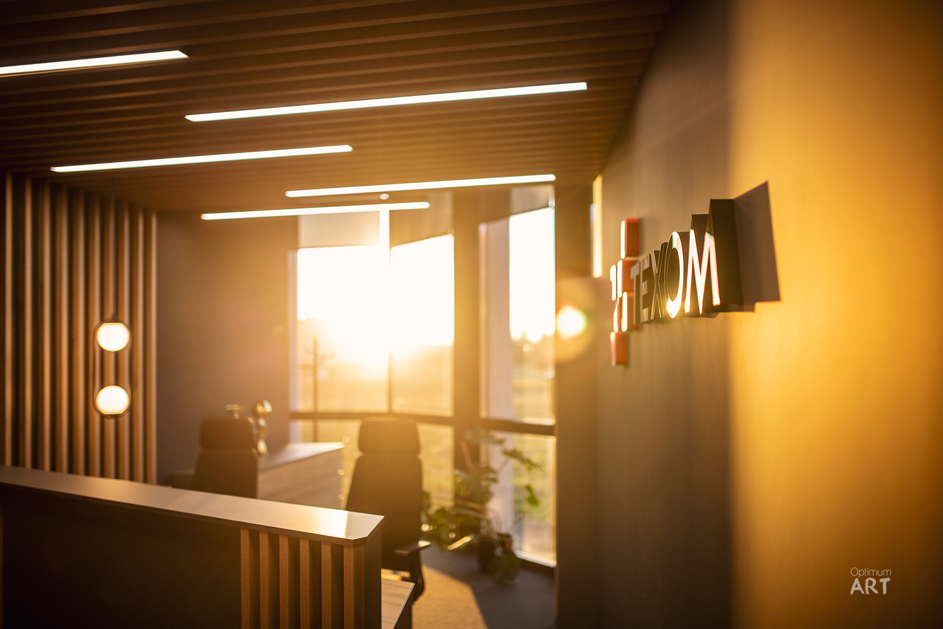 Nowoczesne wnętrze biurowe z drewnianymi panelami na suficie i ścianie, oświetlone ciepłym światłem słonecznym wpadającym przez duże okna. Na pierwszym planie widoczny jest biały blat recepcji, a w tle nowoczesne meble biurowe i dekoracyjne elementy na ścianie.