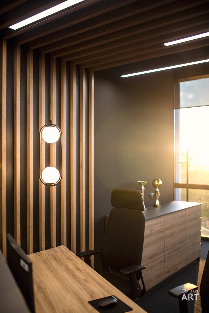 Nowoczesne biuro z drewnianymi lamelami na ścianie, czarnym fotelem przy drewnianym biurku i dwoma okrągłymi lampami. Wnętrze ma ciepłe, naturalne oświetlenie z dużego okna po prawej stronie.