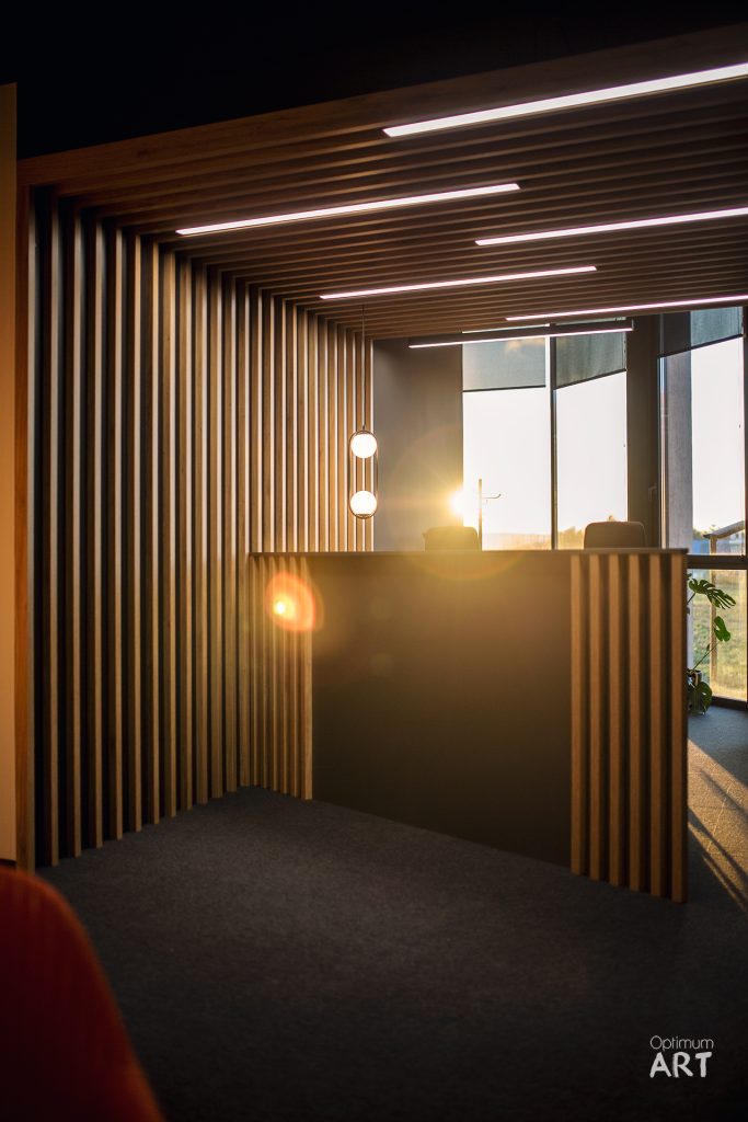 Nowoczesne wnętrze biurowe z drewnianymi lamelami na ścianach i suficie, oświetlone liniowymi lampami LED i okrągłymi lampami wiszącymi. Przez duże okna wpadają promienie słońca, oświetlając przestrzeń z ciemnoszarą wykładziną i czerwonym fotelem w rogu.
