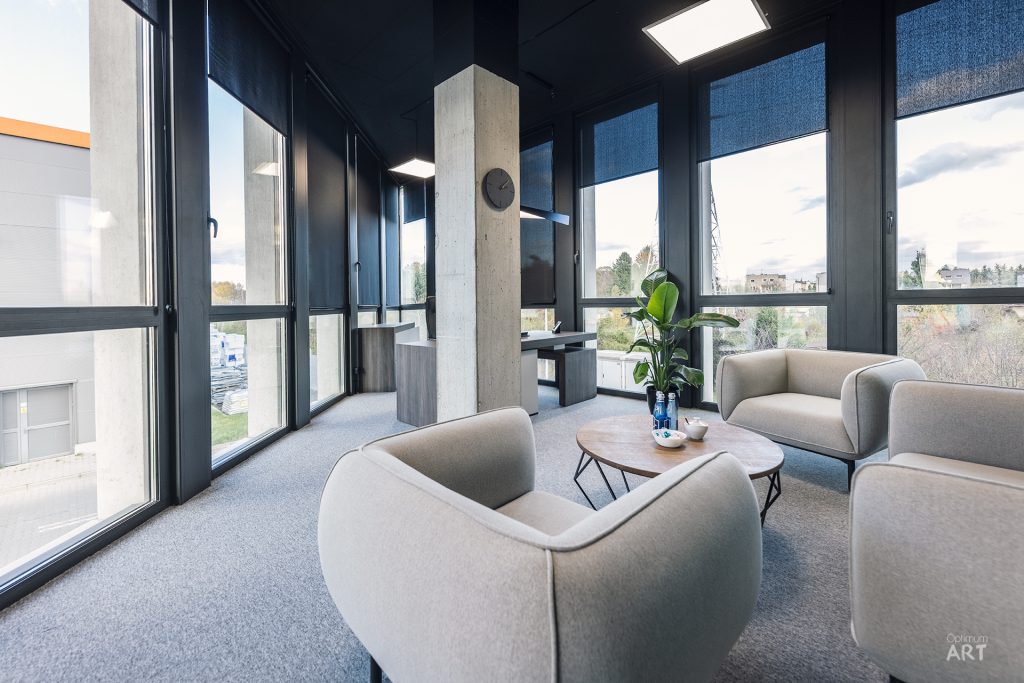 Nowoczesne wnętrze biurowe z dużymi oknami od podłogi do sufitu i szarymi fotelami ustawionymi wokół okrągłego stolika. Pomieszczenie ma ciemne ściany, betonową kolumnę i widok na zewnętrzne budynki przez panoramiczne okna.