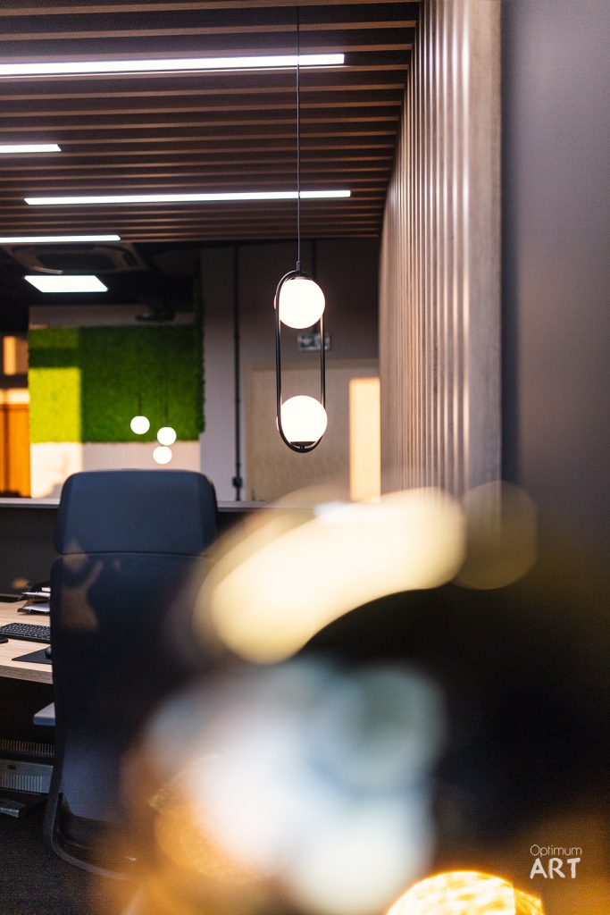 Nowoczesne wnętrze biurowe z drewnianym sufitem z listwami, niebieskim fotelem przy biurku i wiszącą lampą z dwoma okrągłymi kloszami. Na pierwszym planie widoczna jest niewyraźna postać osoby w ruchu.