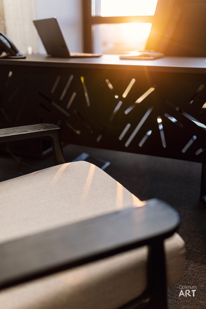 Biurowe krzesło z perforowanym oparciem stoi przy biurku, na którym znajduje się laptop. W tle widać okno z jasnym światłem słonecznym wpadającym do pomieszczenia.