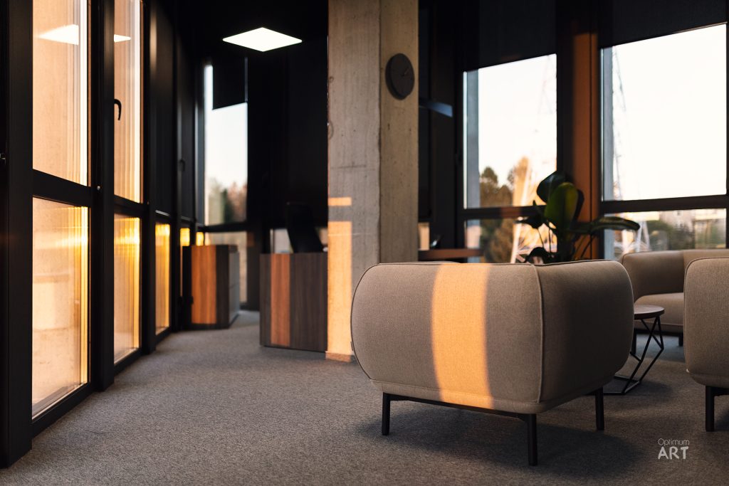 Nowoczesne wnętrze z szarymi fotelami na metalowych nogach, oświetlone ciepłym światłem zachodzącego słońca wpadającym przez duże okna. W tle widoczne są betonowe kolumny i rośliny, a na podłodze leży szary dywan.