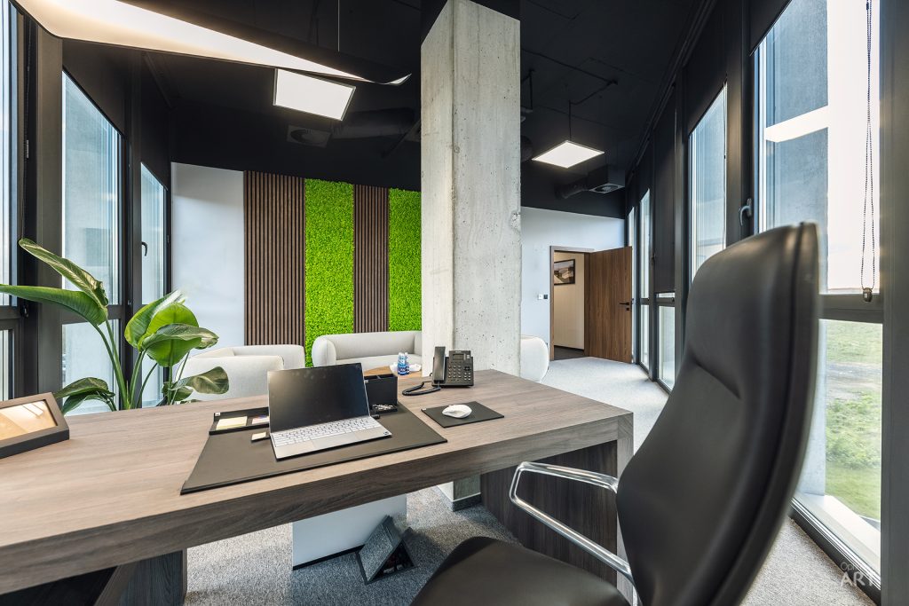 Nowoczesne biuro z drewnianym biurkiem, czarnym fotelem i laptopem, otoczone dużymi oknami. W tle widoczna ściana z zielonymi panelami mchowymi, betonowy słup i jasna strefa wypoczynkowa z białymi fotelami.