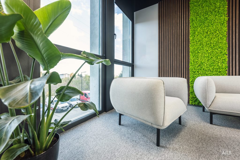 Nowoczesne wnętrze biurowe z dwoma jasnoszarymi fotelami na metalowych nogach, dużą rośliną doniczkową przy oknie i ścianą pokrytą zielonym mchem dekoracyjnym oraz drewnianymi lamelami. Pomieszczenie ma szarą wykładzinę dywanową i duże okna z widokiem na parking.