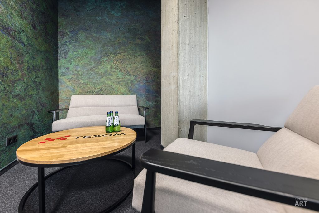 Nowoczesne wnętrze z dwoma beżowymi fotelami z czarnymi metalowymi ramami, ustawionymi wokół okrągłego stolika kawowego z drewnianym blatem. Na ścianie widoczna jest kolorowa tapeta z zielono-niebieskim wzorem przypominającym mapę, a obok betonowy słup.