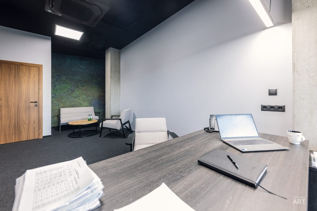 Nowoczesne biuro z dużym drewnianym biurkiem, na którym stoi laptop i leżą dokumenty. W tle widać strefę wypoczynkową z jasnymi fotelami, ciemnoszarą ścianą z zielonym akcentem i drewnianymi drzwiami.
