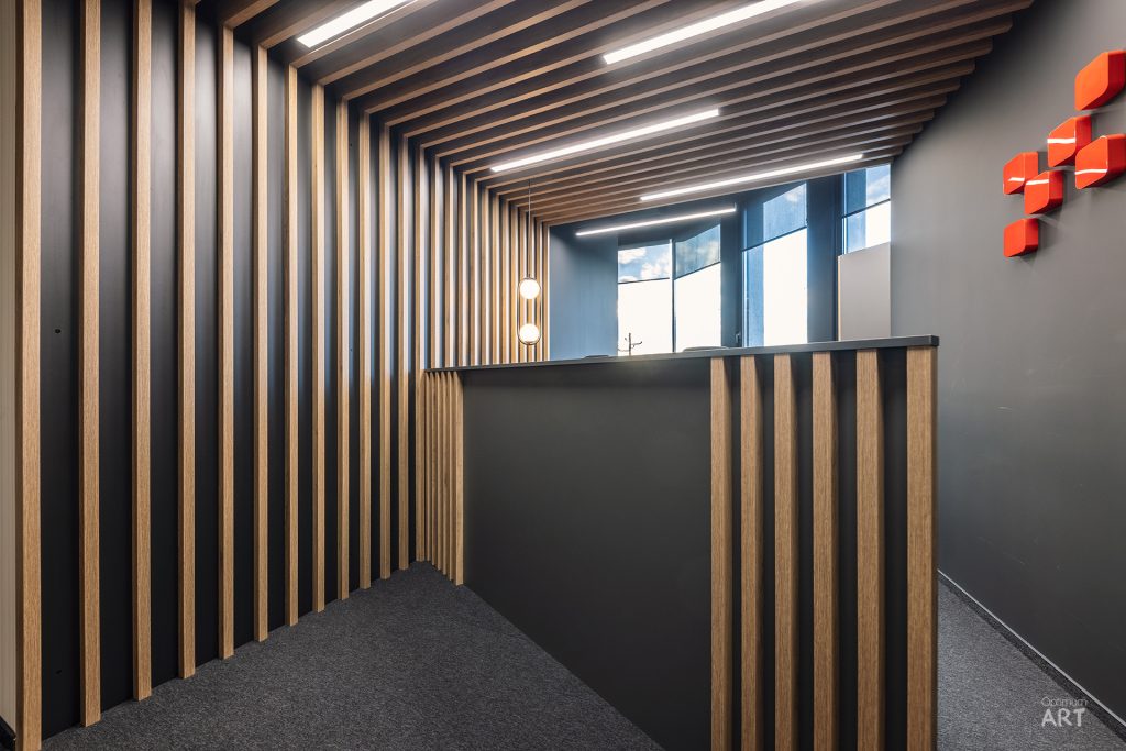 Nowoczesne wnętrze biurowe z drewnianymi lamelami na ścianie i suficie, szarymi ścianami oraz czerwonymi elementami dekoracyjnymi. W głębi widoczne są duże okna wpuszczające naturalne światło oraz nowoczesne oświetlenie sufitowe.