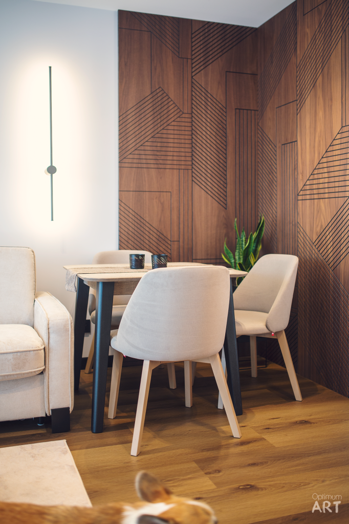 Nowoczesna jadalnia z okrągłym stołem, dwoma beżowymi krzesłami i dekoracyjną drewnianą ścianą z geometrycznym wzorem. W tle widoczny jest fragment beżowego fotela i minimalistyczna lampa ścienna.