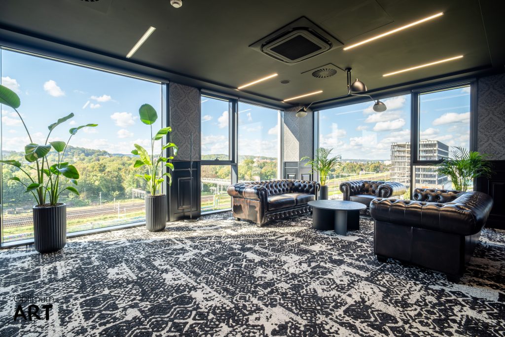 Nowoczesne biurowe pomieszczenie wypoczynkowe z trzema skórzanymi fotelami Chesterfield ustawionymi wokół okrągłego stolika na wzorzystym dywanie. Przestrzeń otoczona jest panoramicznymi oknami z widokiem na miasto i zieleń, a w rogu stoją duże rośliny doniczkowe.