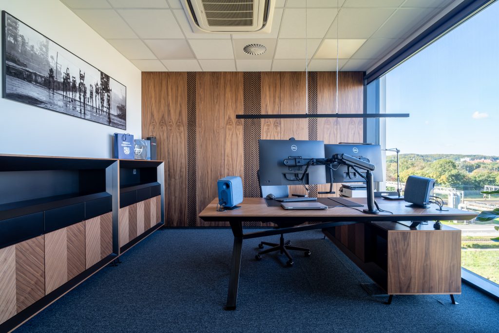 Nowoczesne biuro z drewnianym biurkiem, dwoma monitorami komputerowymi i ergonomicznym krzesłem obrotowym. Pomieszczenie ma drewniane wykończenia ścian, duże okno z widokiem na zieleń oraz niebieską wykładzinę dywanową.