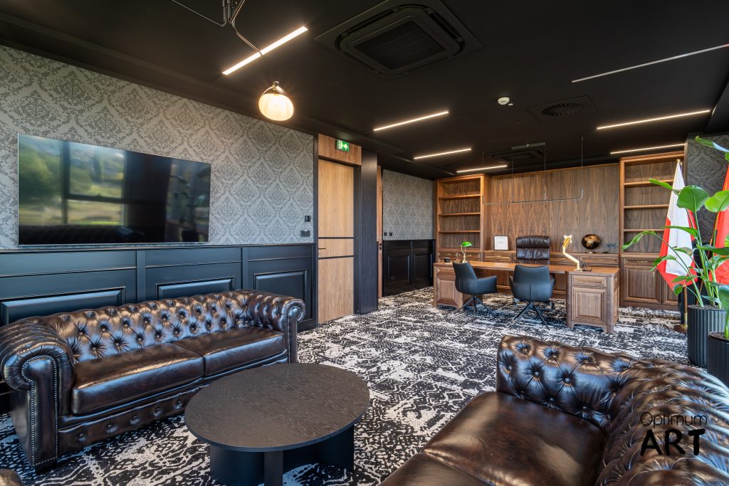 Luksusowe biuro z ciemnymi skórzanymi fotelami Chesterfield, drewnianym biurkiem i telewizorem na ścianie. Wnętrze ma elegancki design z czarnym sufitem, wzorzystymi tapetami i drewnianymi meblami.
