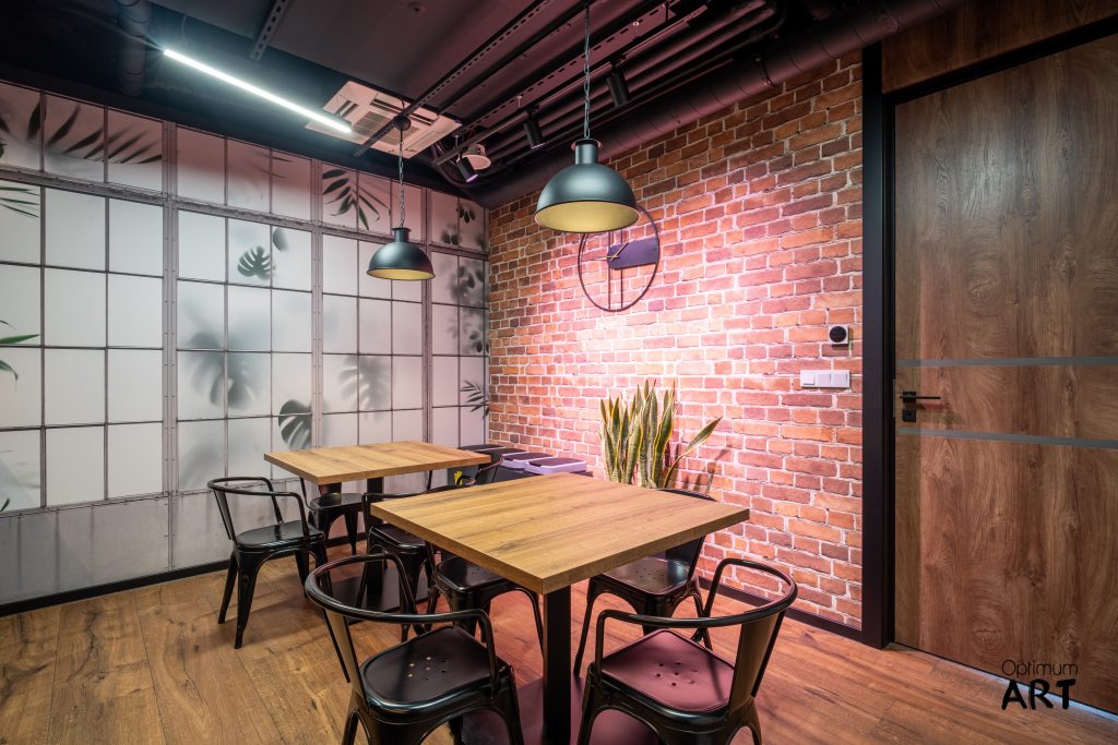 Wnętrze nowoczesnej kawiarni lub restauracji w stylu industrialnym z ceglaną ścianą, drewnianymi stołami, czarnymi krzesłami metalowymi i lampami wiszącymi. Po lewej stronie znajduje się przeszklona ściana z roślinnymi motywami, a po prawej drewniane drzwi.