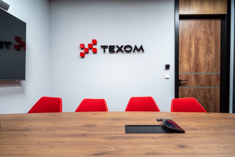 Sala konferencyjna z drewnianym stołem i czterema czerwonymi krzesłami, na białej ścianie widnieje logo "TEXOM" z czerwonymi elementami geometrycznymi. W tle znajdują się drewniane drzwi i monitor na ścianie.