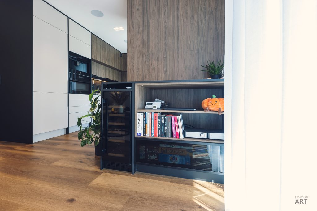 Nowoczesna kuchnia z drewnianymi i białymi szafkami, w której widać otwarte półki z książkami i dekoracjami. Na podłodze z drewna stoją rośliny doniczkowe, a w tle znajdują się wbudowane urządzenia AGD.