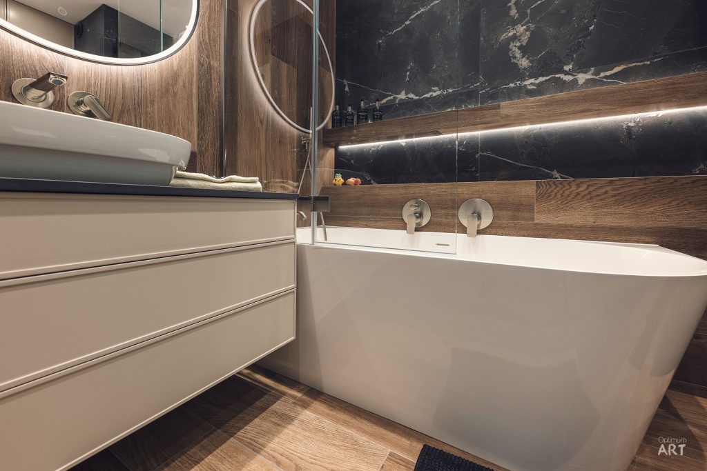 Nowoczesna łazienka z białą wolnostojącą wanną, drewnianymi elementami wykończeniowymi i ciemnoszarym marmurem na ścianie. Po lewej stronie znajduje się biała szafka pod umywalkę z okrągłym lustrem i LED-owym oświetleniem.