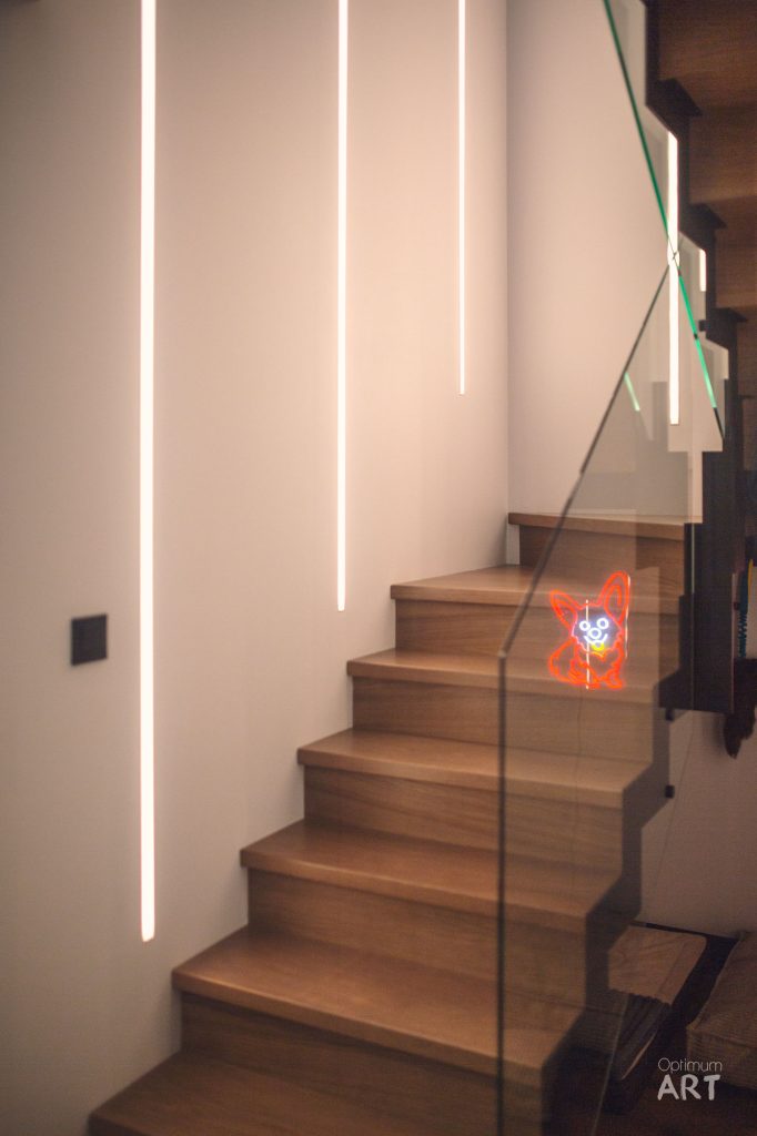 Nowoczesne drewniane schody z szklaną balustradą prowadzą w górę wzdłuż białej ściany oświetlonej pionowymi pasami LED. Na jednym ze stopni widoczna jest czerwona dekoracja świetlna w kształcie kwiatu.